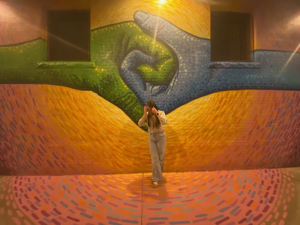 Woman standing in front of a yellow mural with a green and blue hand holding each other.