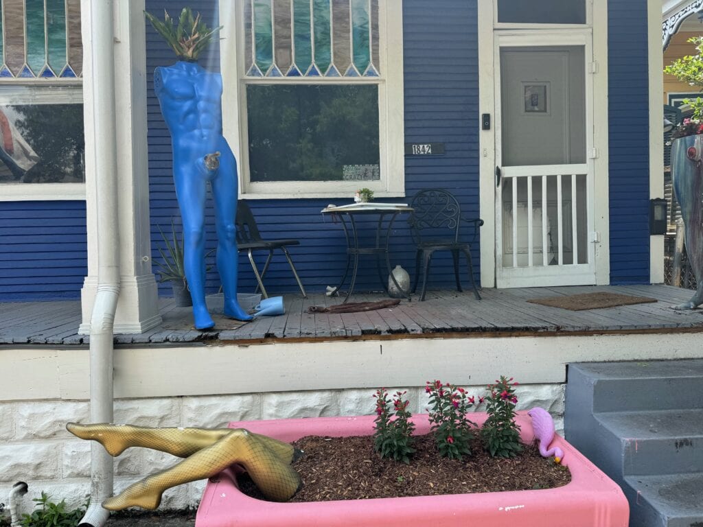 A hilarious porch in New Orleans. In front is a pair of fishnet-clad legs poking out of a flower bed. Behind is a statue of a man with plants for a head and penis.