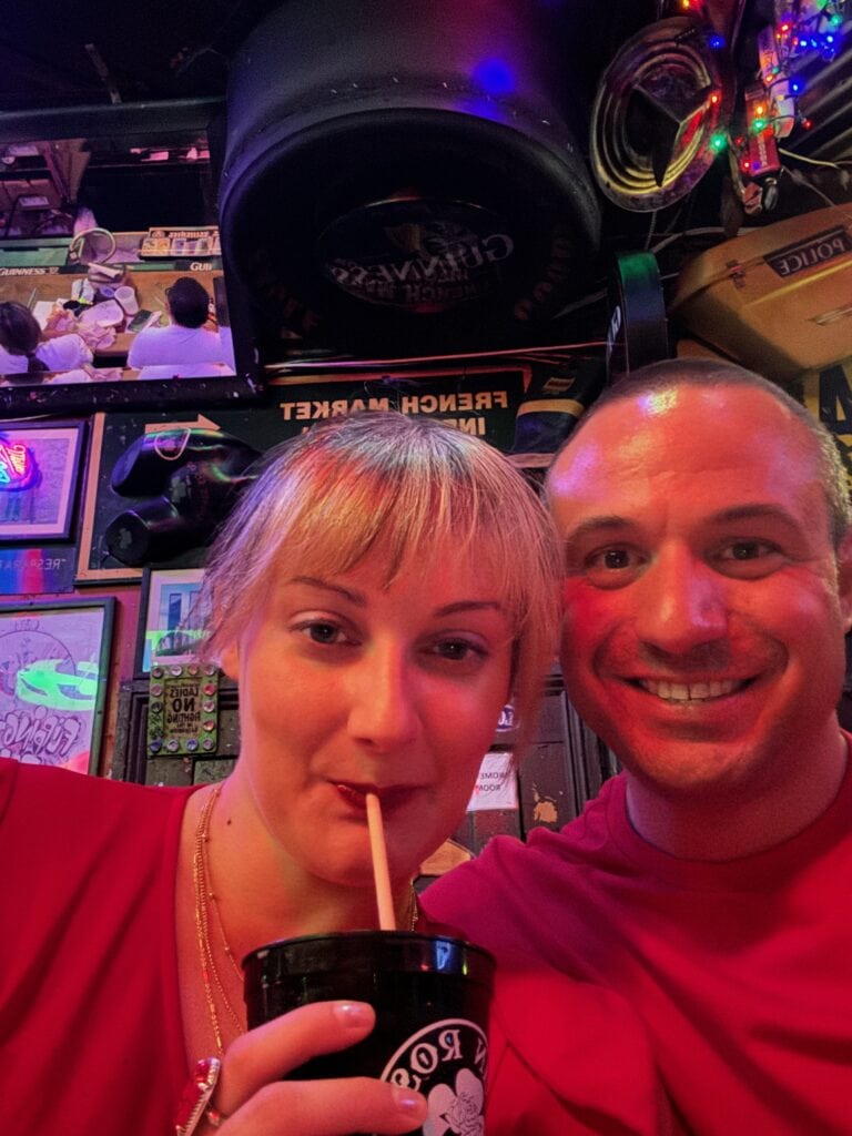 woman and man wearing red posing in front of bar signs