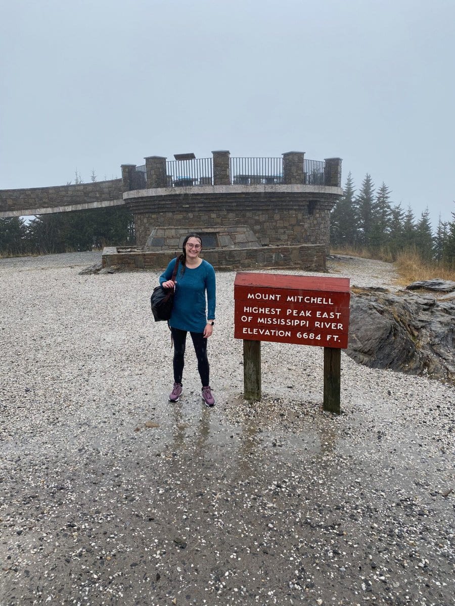 Woman drenched in rain water posing by Mt. Mitchell sign at the summit of the trail.