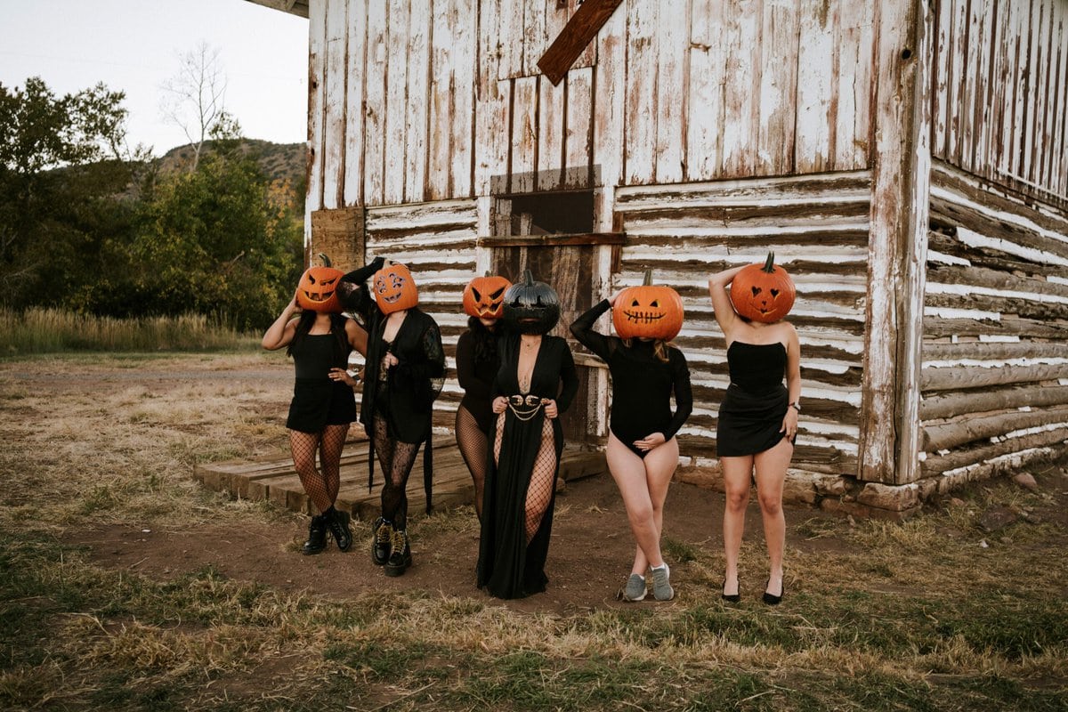 Five people wearing black outfits and pumpkin masks stand in front of a rustic wooden building outdoors, posing together on a patch of grass and dirt.