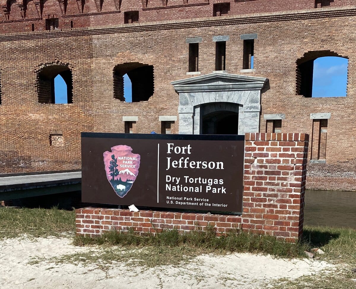 A sign for Fort Jefferson at Dry Tortugas National Park stands in front of a brick fort wall with arched windows and a stone entrance, under a sunny blue sky—one of many reasons to visit the Florida Keys.