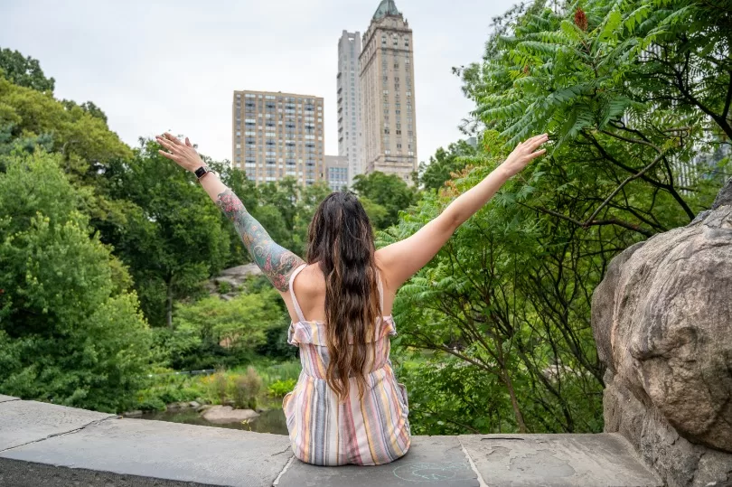 Back of woman sitting in Central Park with arms raised in a V shape