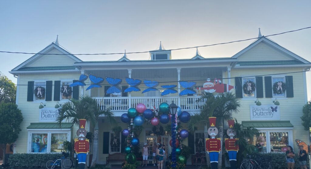 A yellow, two-story building decorated with colorful balloons, blue butterfly decorations, and two large toy soldier figures by the entrance. This lively spot is perfect for anyone searching for fun things to do in Key West. Palm trees shade people near the door.