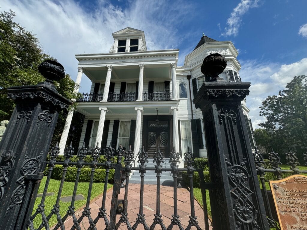 A tall, historic white mansion with columns and balconies stands behind a decorative black iron gate under a blue sky with scattered clouds—a must-see for those seeking things to do alone in New Orleans. A plaque is visible near the entrance.