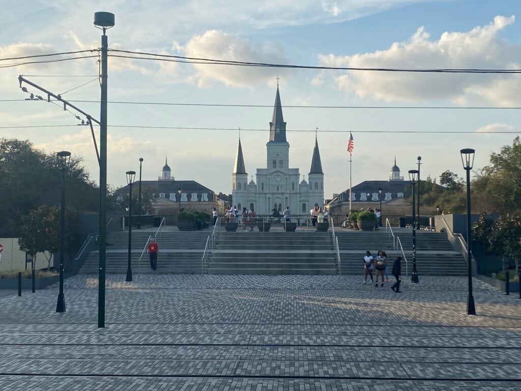 St. Louis Cathedral in New Orleans stands behind a wide staircase and open plaza, where people gather under a partly cloudy sky. Streetcar tracks and lamp posts line the foreground, making it a perfect spot for those seeking things to do alone in New Orleans.