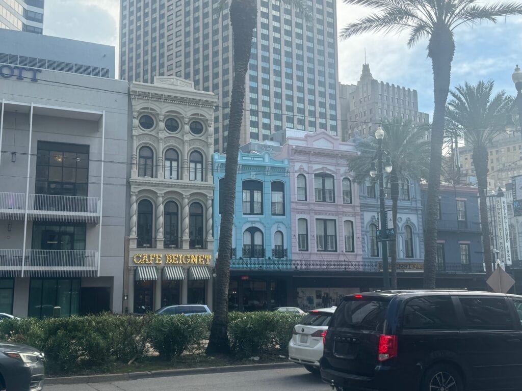 Colorful historic buildings, including a blue and pink one housing Café Beignet, line a street in New Orleans—a great spot for those seeking things to do alone in New Orleans. Palm trees and parked cars sit below a modern skyscraper.