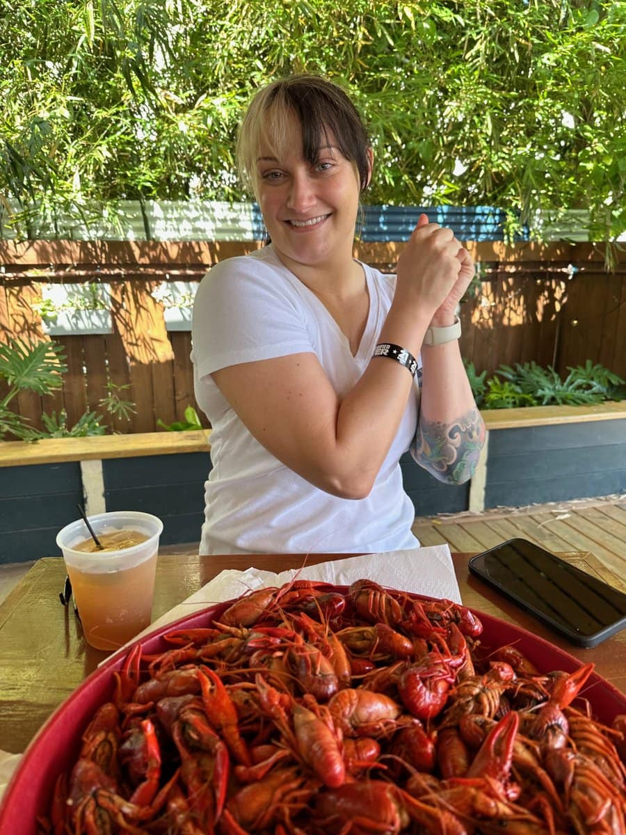 A smiling person in a white shirt sits at an outdoor table with a large red tray of boiled crawfish and a drink—one of the best things to do alone in New Orleans—surrounded by greenery and wooden fencing.
