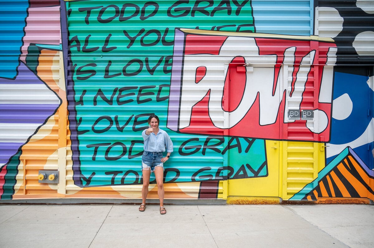 Woman holding fist out, pretending to punch the air while standing next to a mural that says 