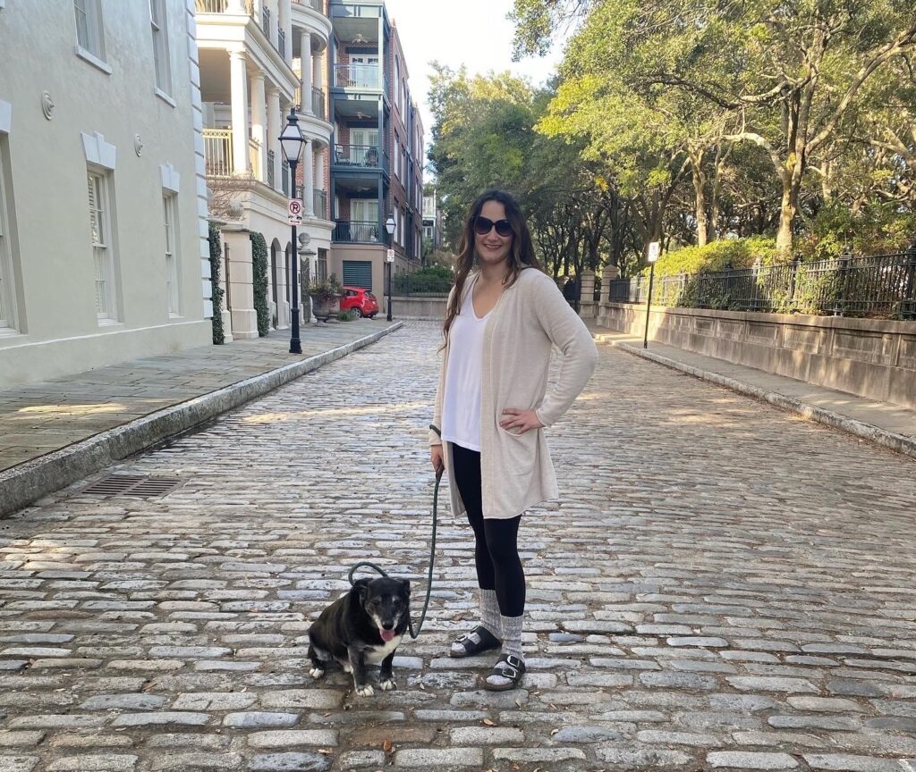 A digital nomad with a dog, wearing sunglasses and a light sweater, stands on a cobblestone street. Trees and historic buildings line the background as she holds the leash of her small black and white companion.