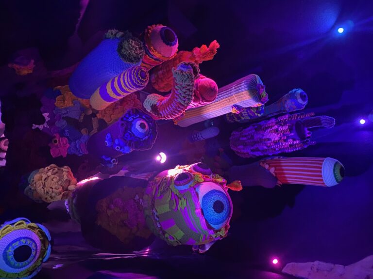 Colorful, crocheted sculptures resembling abstract sea creatures and coral are illuminated by purple, pink, and blue lights in a dark room—a vibrant, surreal underwater effect perfect for those seeking unique things to do in Denver alone.