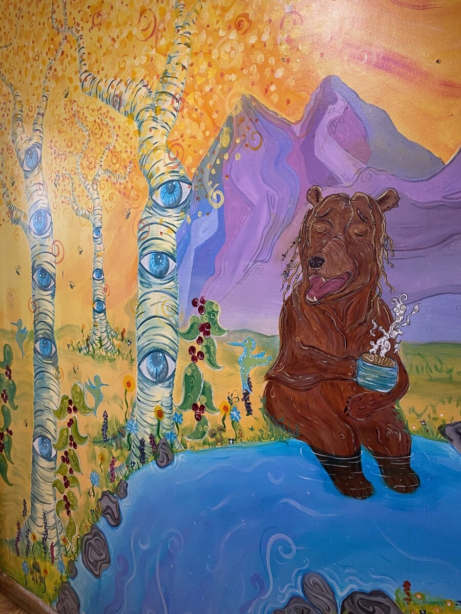 A whimsical mural in Idaho Springs features a bear with dreadlocks by a blue stream, holding a steaming mug. Birch trees with eyes, colorful flowers, and purple mountains fill the surreal scene—one of the unique things to do in Idaho Springs, CO.