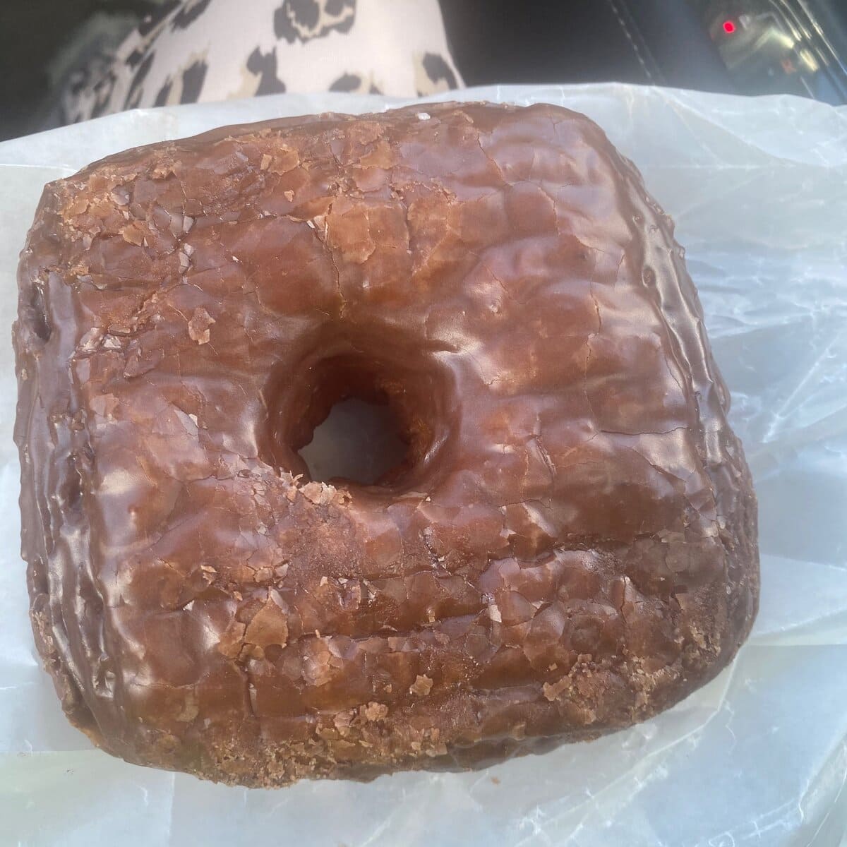 A square-shaped glazed donut with a hole in the center sits on a piece of wax paper, with a persons lap and patterned clothing visible in the background.