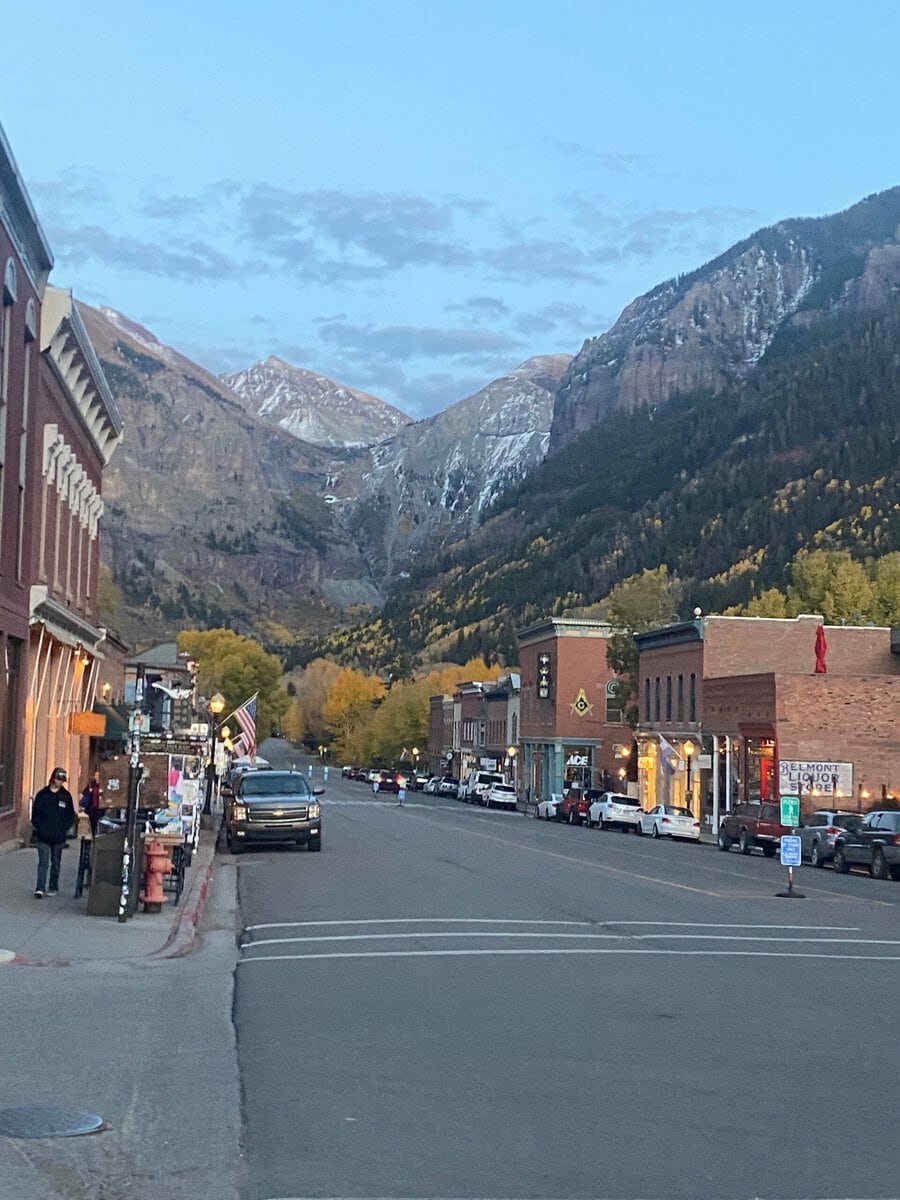 A small mountain town street at dusk, lined with brick buildings and cars, with people strolling the sidewalks—captures the charm and things to do in Idaho Springs, CO. Snow-capped peaks and autumn trees rise under a partly cloudy sky.