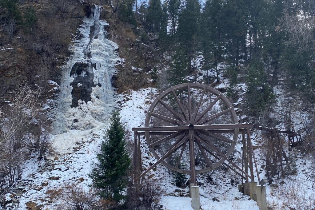 A large wooden water wheel stands in front of a partially frozen waterfall on a rocky, snow-dusted hillside surrounded by evergreen and bare trees.