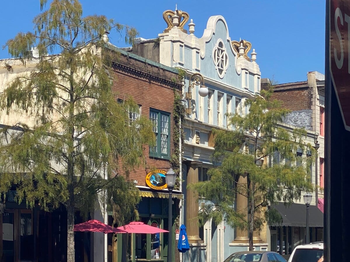 A city street features historic buildings with ornate facades, trees lining the sidewalk, and pink umbrellas shading outdoor seating in front of a restaurant on a sunny day.