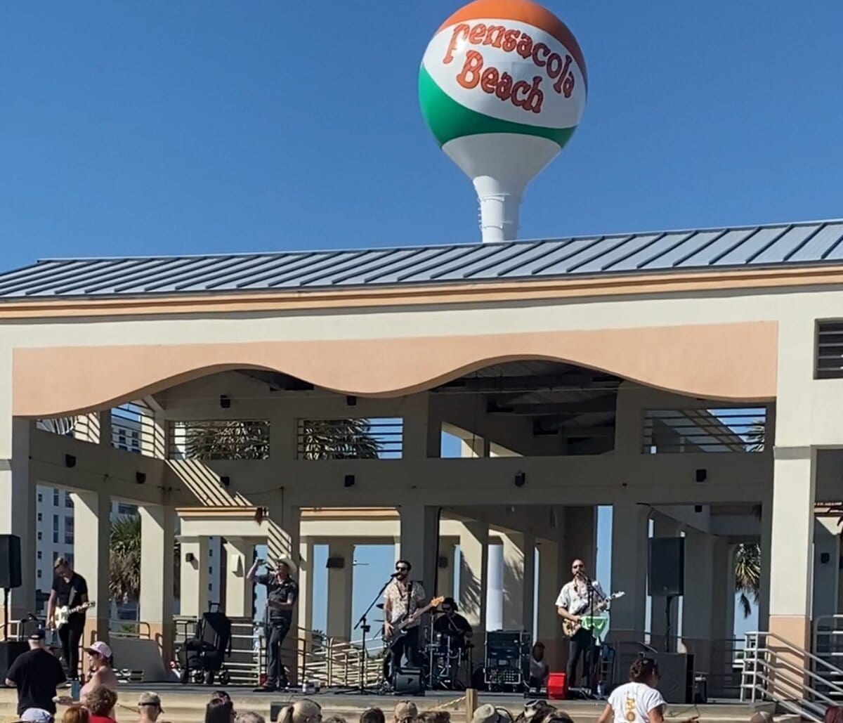 A band rocks an outdoor stage under a clear blue sky, as a beach-ball water tower marked Pensacola Beach stands behind them. Fans—many on solo travel exploring Alabama's Gulf Coast—gather to enjoy the lively show.