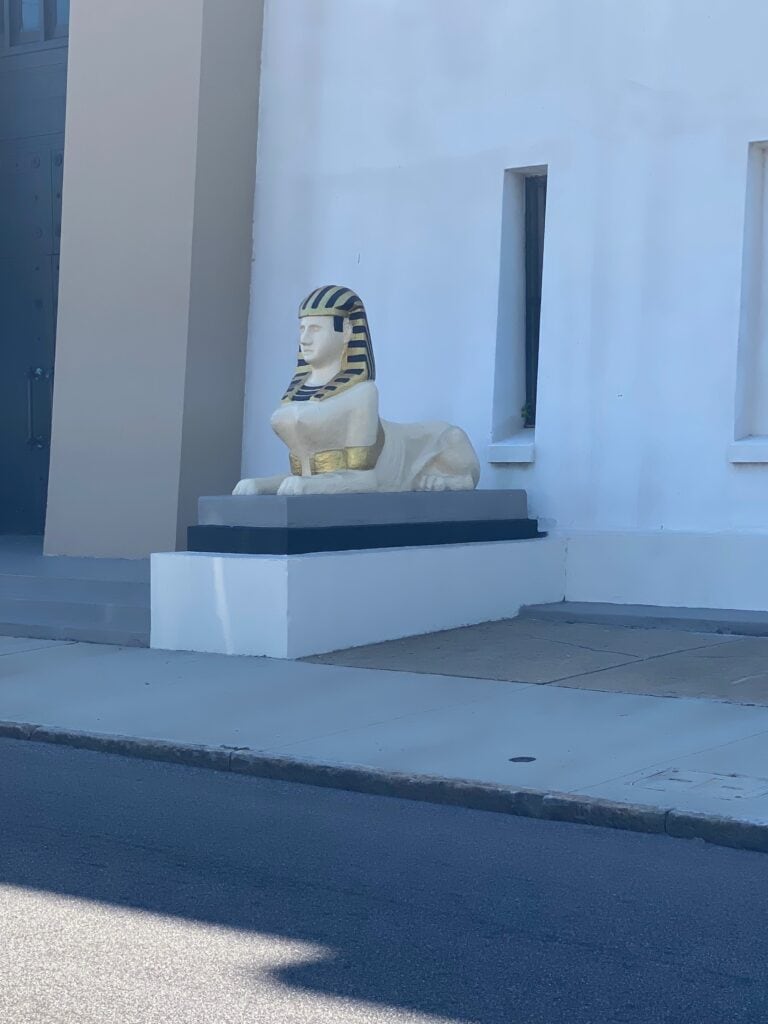 A small sphinx statue with a gold and black striped headdress sits on a white platform outside a light-colored building—an unexpected find for those enjoying solo travel along Alabama's Gulf Coast.
