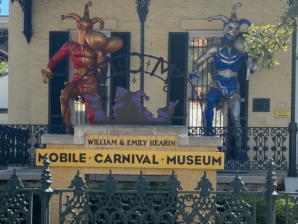 Two large jester statues, one red and one blue, stand on a balcony above a sign reading Mobile Carnival Museum William & Emily Hearin on a yellow brick building.