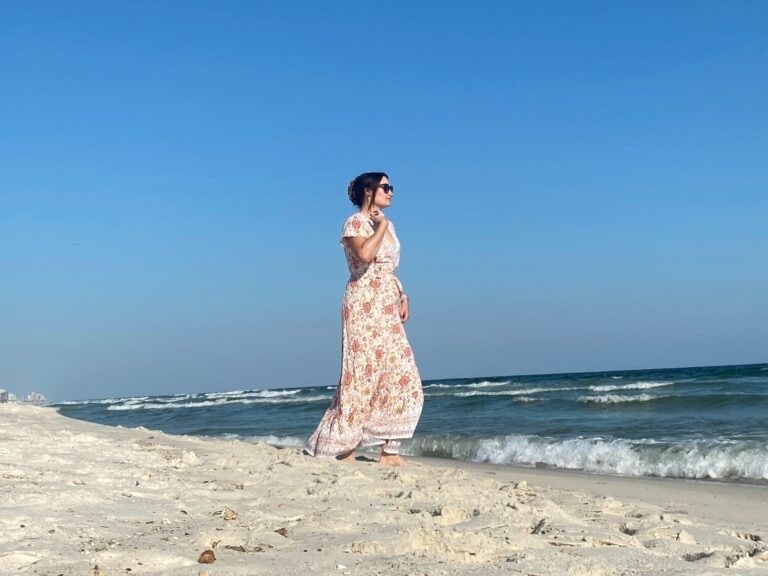 A woman wearing a long floral dress and sunglasses walks along a sandy beach near the ocean under a clear blue sky. Waves are gently rolling in, and the shoreline extends into the distance.