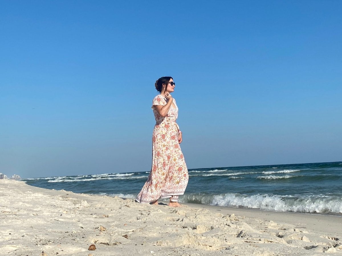 A woman wearing a long floral dress and sunglasses walks along a sandy beach near the ocean under a clear blue sky. Waves are gently rolling in, and the shoreline extends into the distance.