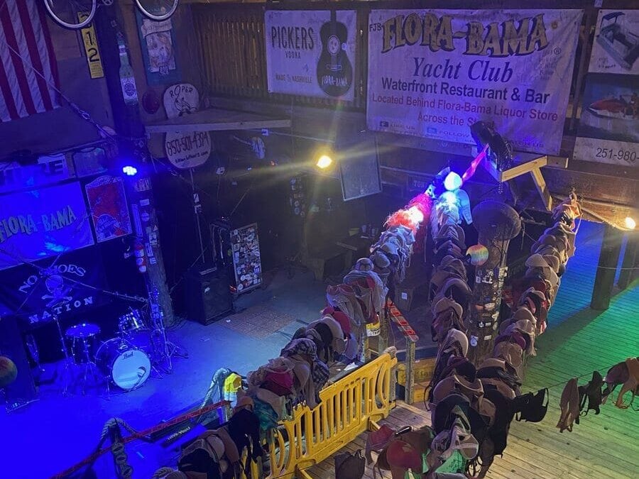A dimly lit bar interior with a stage and drum set in one corner. Numerous bras hang along a railing, and banners advertising Flora-Bama Yacht Club and other venues are displayed on the walls.