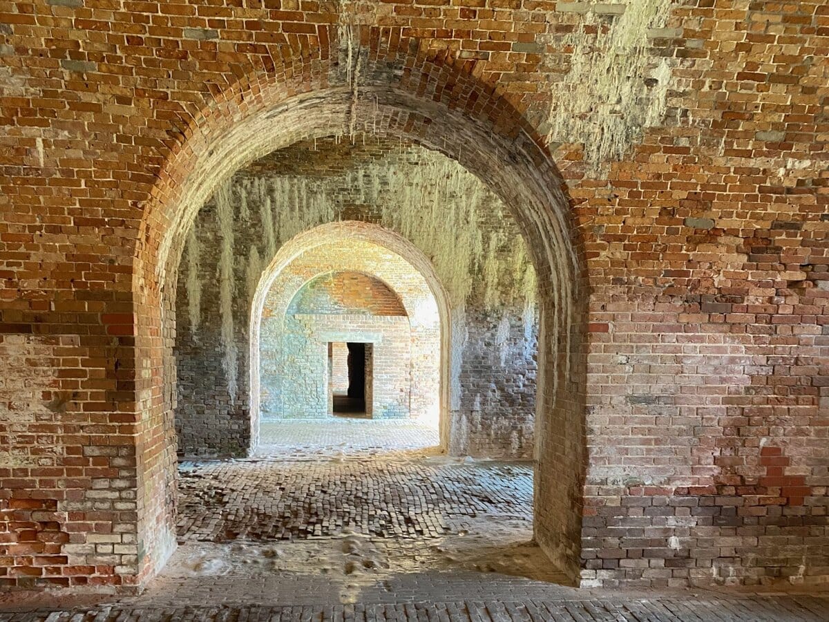 View of a brick-walled arched hallway with multiple archways receding into the distance, leading to a doorway with sunlight illuminating the textured brick floors and walls.