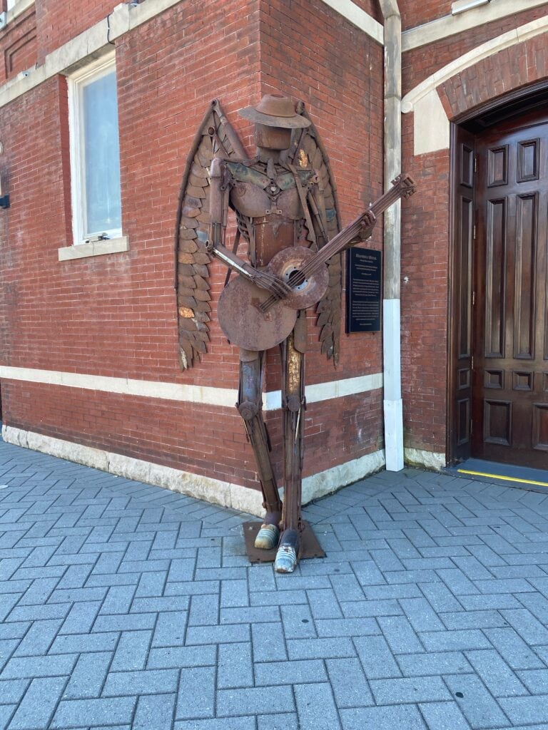 A metal sculpture of an angel with wings, wearing a hat and playing a guitar, stands on a brick sidewalk by a red brick building with a wooden door—an inspiring find for solo travel along Alabama's Gulf Coast.