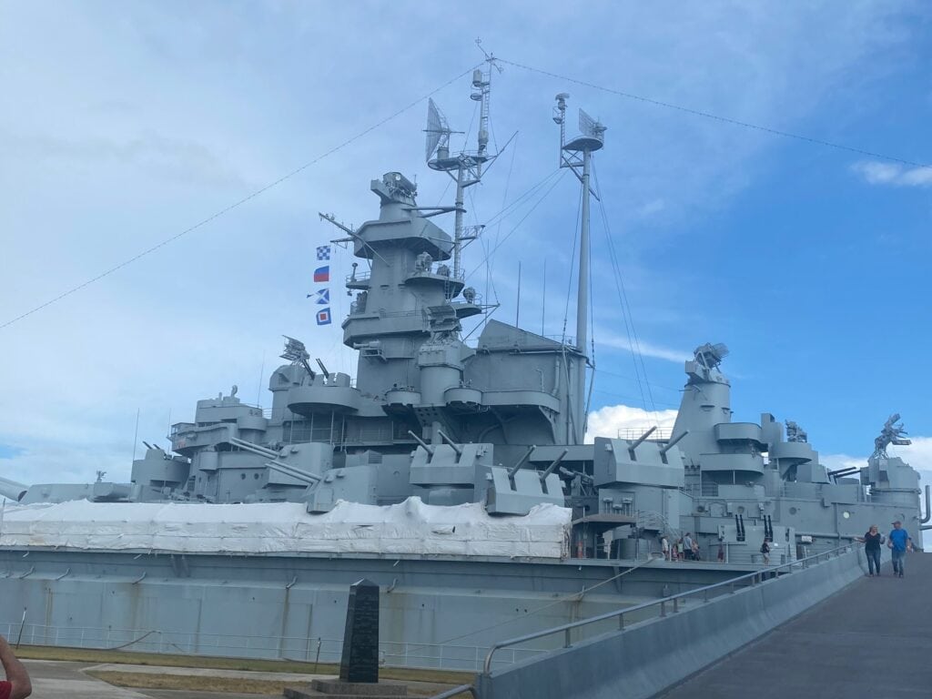 A large grey battleship with radar towers and signal flags is docked under a partly cloudy sky. Several people are walking nearby on a paved path leading up to the ship.