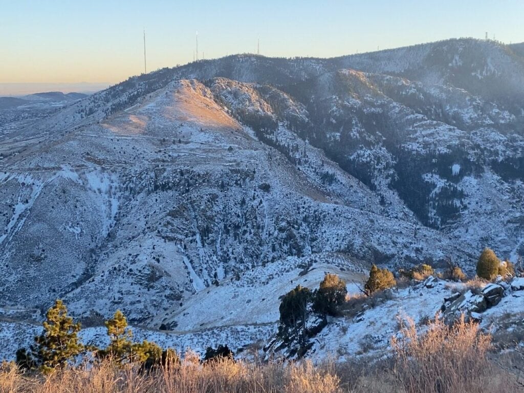 Snow-dusted mountains under a clear sky at sunrise, with sunlight casting a warm glow on the peaks. Sparse trees and brush dot the rocky slopes, creating a tranquil winter landscape.