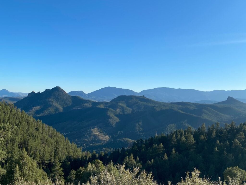 A scenic view of forested mountains under a clear blue sky, with layers of green hills and distant peaks stretching across the horizon.