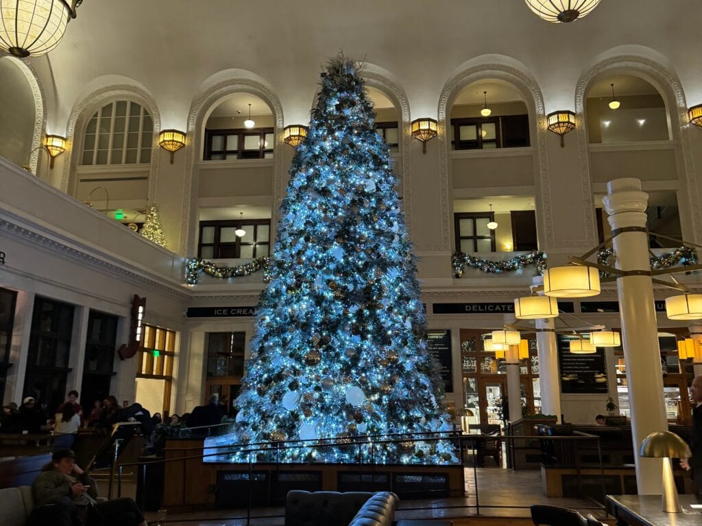 A large, beautifully decorated Christmas tree with blue and white lights stands in the center of a grand, softly lit indoor hall with arched windows and festive garlands. People sit and relax around the tree.