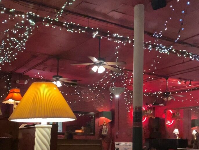 A dimly lit restaurant with red walls and ceiling, string lights, ceiling fans, glowing lamps, and neon signs creating a cozy, eclectic atmosphere.