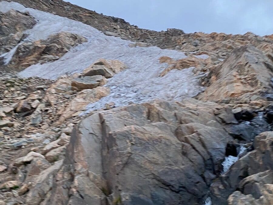 Rocky mountain slope with patches of ice and snow, featuring large boulders and rough terrain under a cloudy sky.