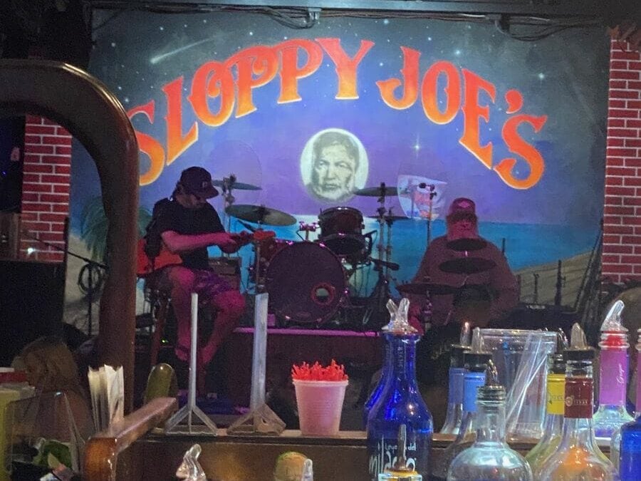 Two musicians play drums on a stage with a “Sloppy Joe’s” mural behind them, featuring a large face and a beach scene. Bottles and bar items are in the foreground. The setting appears to be a lively bar or club.