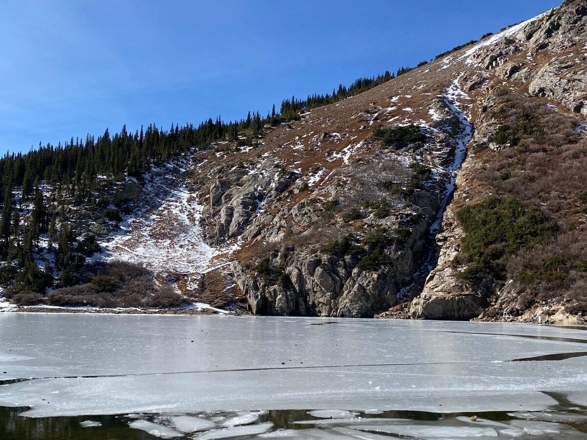 A partially frozen lake sits at the base of a rocky, snow-dusted hill covered with evergreen trees under a clear blue sky.
