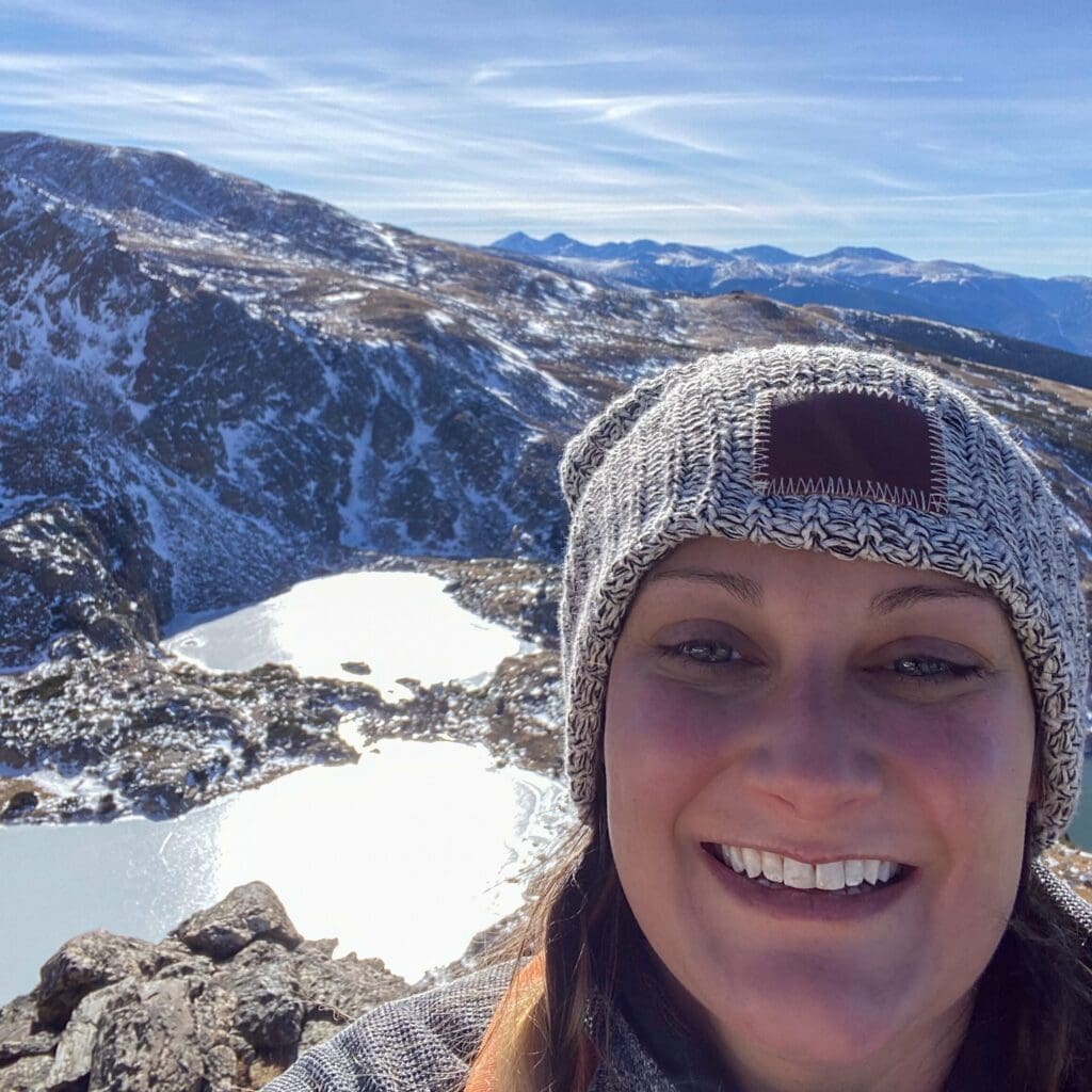 A woman wearing a knit hat smiles in the foreground with snow-covered mountains and frozen lakes in the background under a clear blue sky.