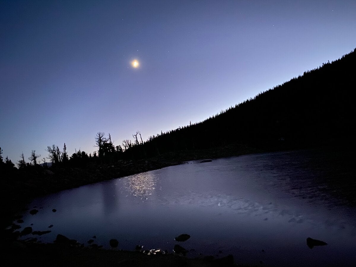 A serene twilight scene shows a calm lake beneath a dark hill silhouette, with a glowing moon and fading blue sky, reflected gently on the water—reminiscent of views along the James Peak Trail. Sparse trees line the ridge above the lake.