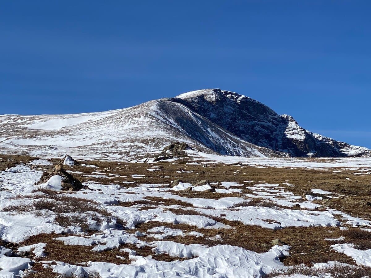 A snow-dusted mountain peak rises under a clear blue sky, with patches of snow and exposed rocky ground in the foreground. The landscape appears rugged and open, with minimal vegetation.