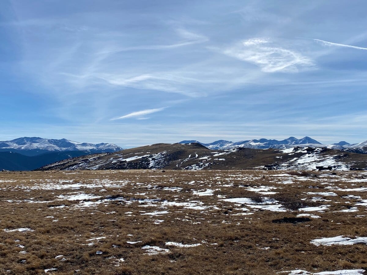 A wide, open landscape with sparse snow patches on grassy, rolling hills, reminiscent of the scenery along the St. Mary's Glacier hike. Snow-capped mountains rise in the background under a partly cloudy blue sky.