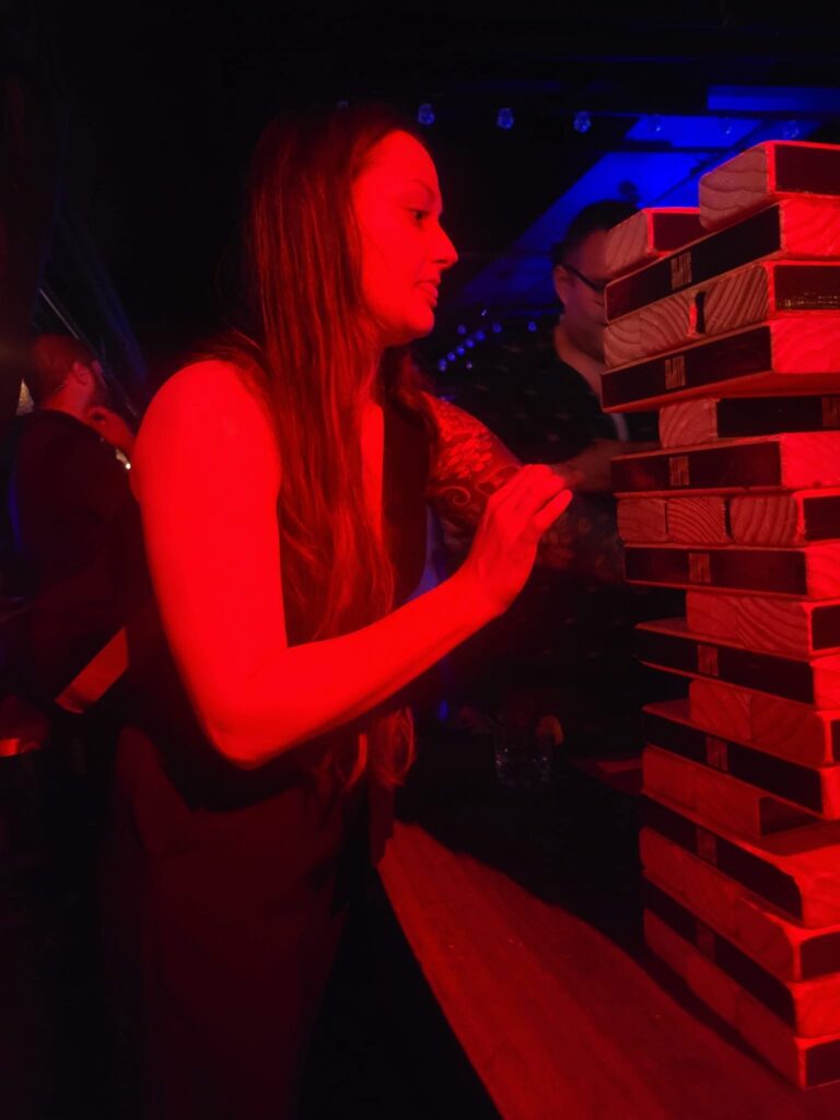 A woman bathed in red light concentrates as she carefully removes a wooden block from a tall Jenga tower—one of the best things to do alone in NYC—at a dimly lit venue.