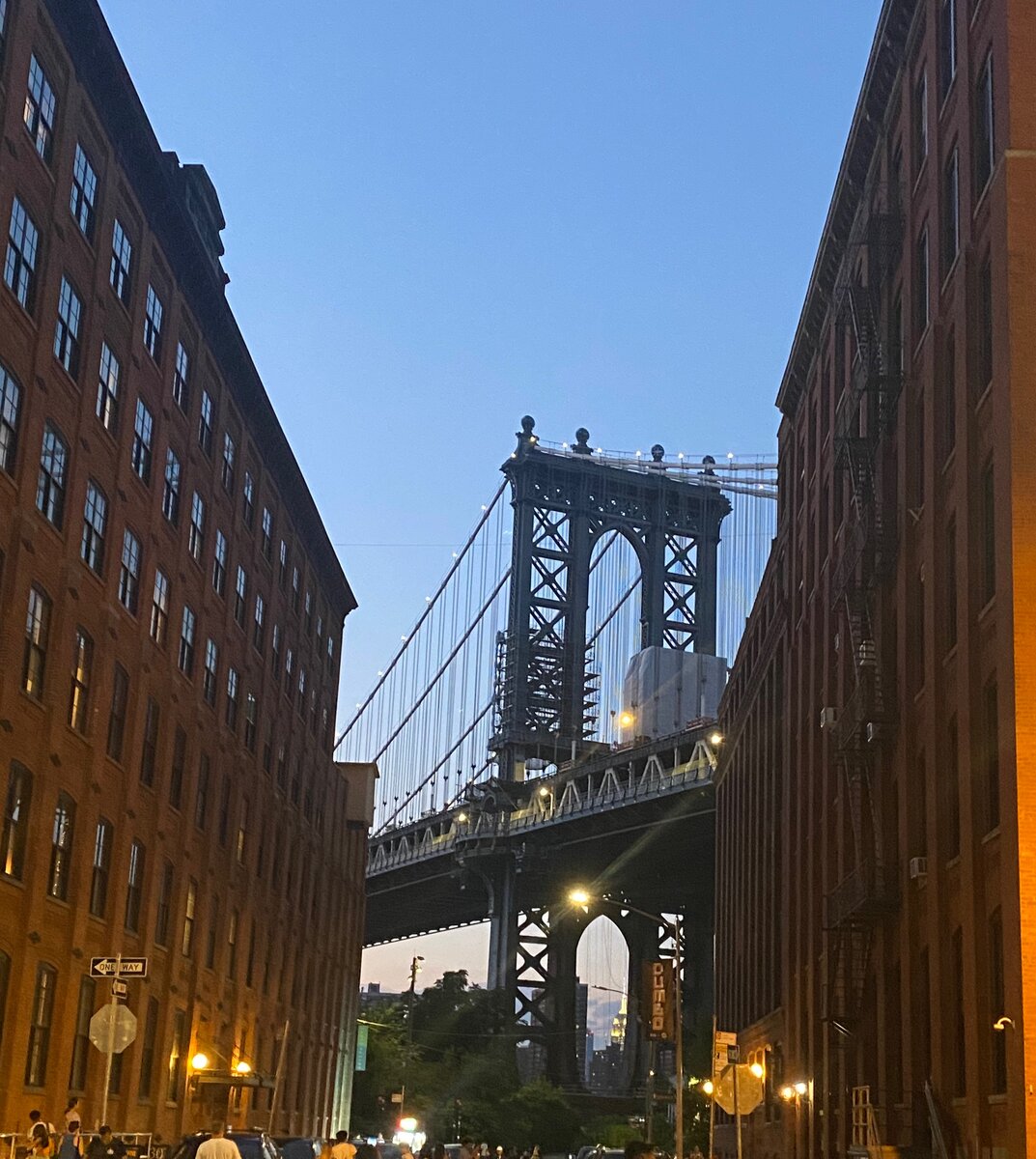 The Manhattan Bridge is framed between two tall brick buildings at dusk, with a clear blue sky in the background. Perfect for SOLO FEMALE travel in NYC, streetlights illuminate the inviting scene below.