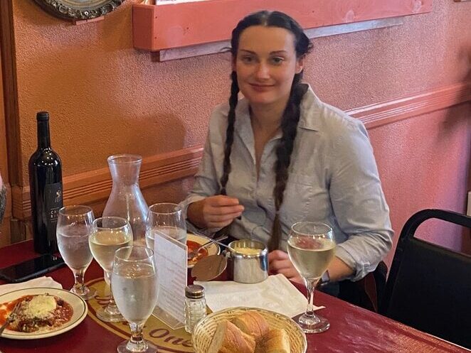 A woman with long brown braids sits at a restaurant table set with plates of food, glasses of water, a bottle of wine, and a basket of bread. She is smiling and wearing a light blue shirt.