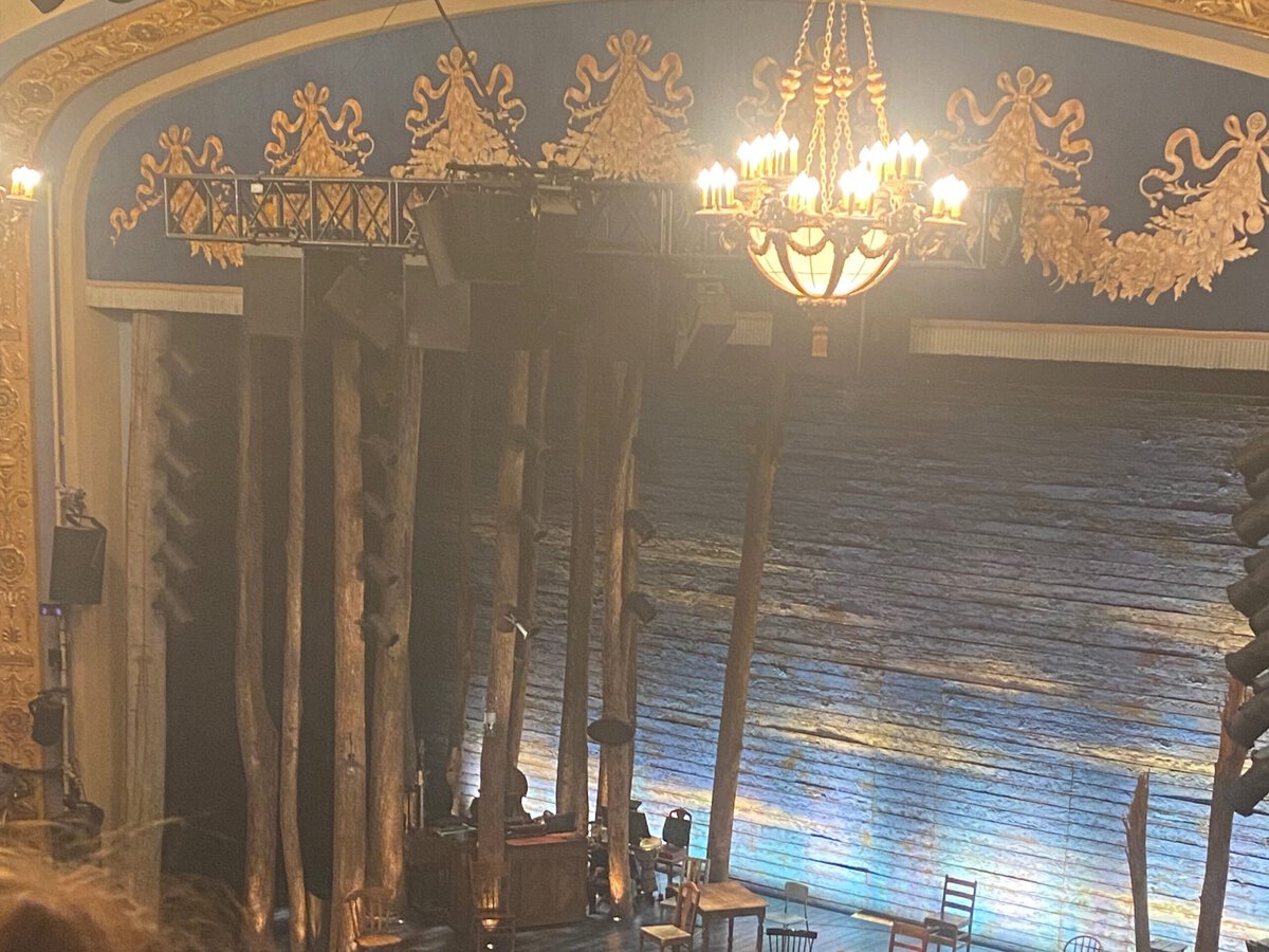 A theater stage set features tall wooden poles, simple furniture, and a painted backdrop resembling a sky. An ornate chandelier and decorative gold trim adorn the ceiling above the stage.