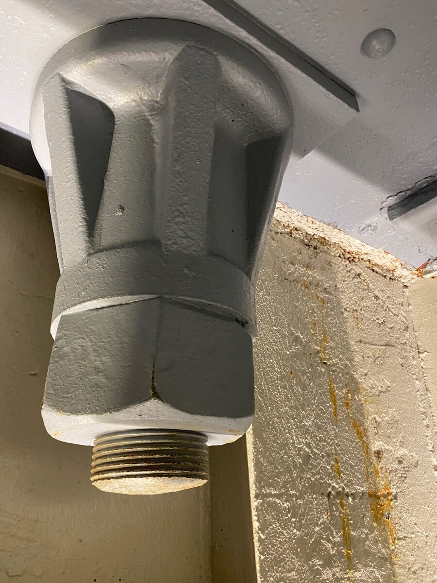 A large, gray, industrial valve or fitting is mounted upside-down on a ceiling, its threaded metal end pointing downward near a wall with peeling paint and rust stains—a gritty scene capturing the hidden side of SOLO FEMALE travel NYC.