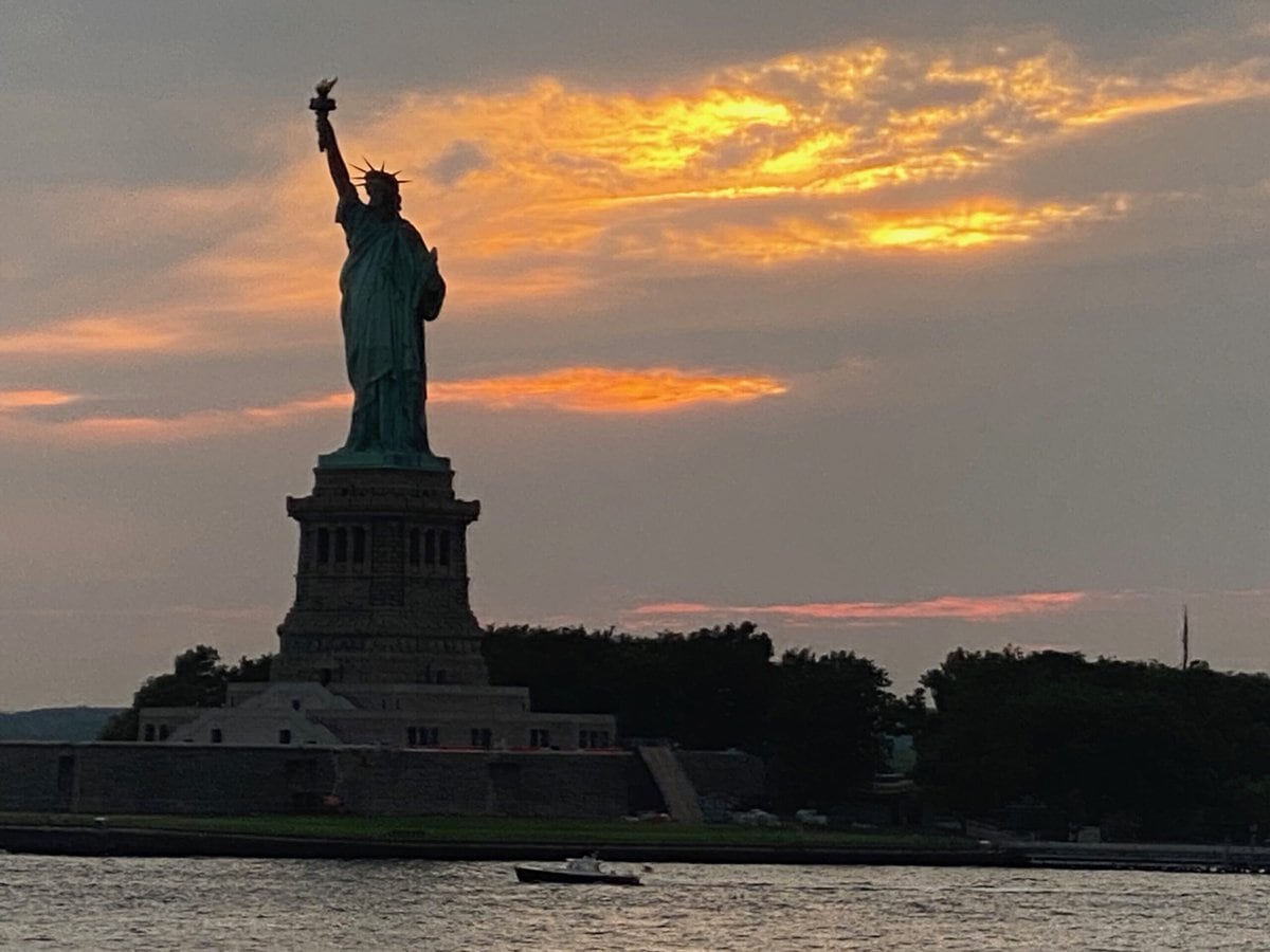 The Statue of Liberty is silhouetted against a dramatic sunset sky with orange clouds, as seen from across the water. Trees and part of the island are visible in the foreground.