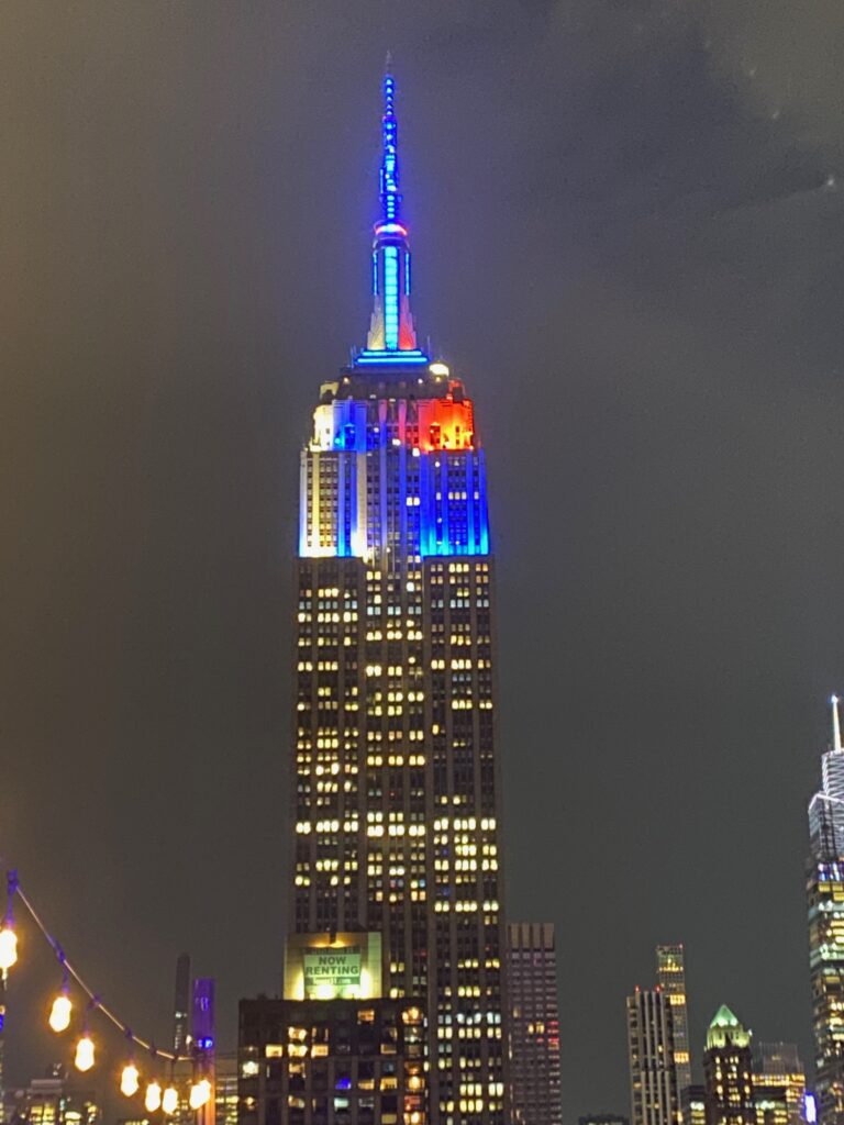The Empire State Building at night, a must-see for SOLO FEMALE travel in NYC, shines with red, white, and blue lights atop its spire against a cloudy sky, surrounded by the city's buildings at its base.