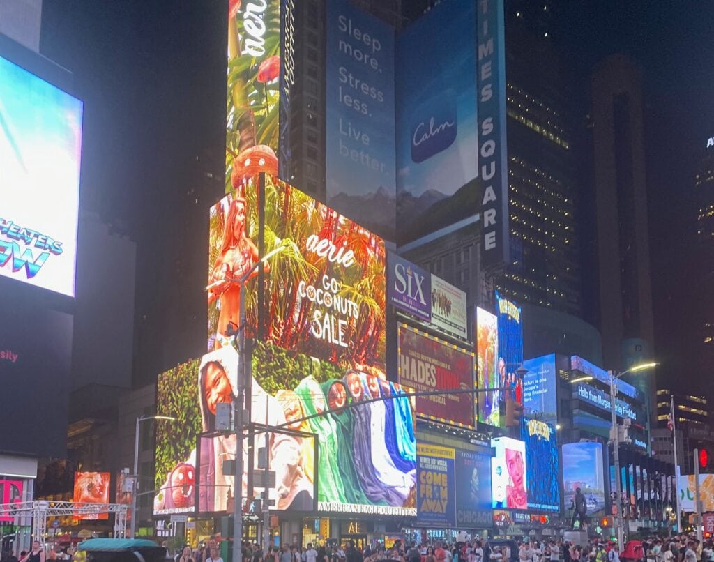 Bright, colorful electronic billboards and advertisements light up Times Square at night, featuring brands like Aerie and Calm. Crowds—and solo female travel enthusiasts—gather on the busy street below the illuminated displays.