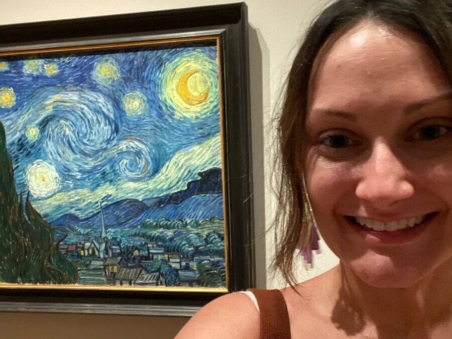 A woman smiles for a selfie in front of Vincent van Gogh’s famous painting The Starry Night displayed in a museum. The painting is visible in the background to her left.