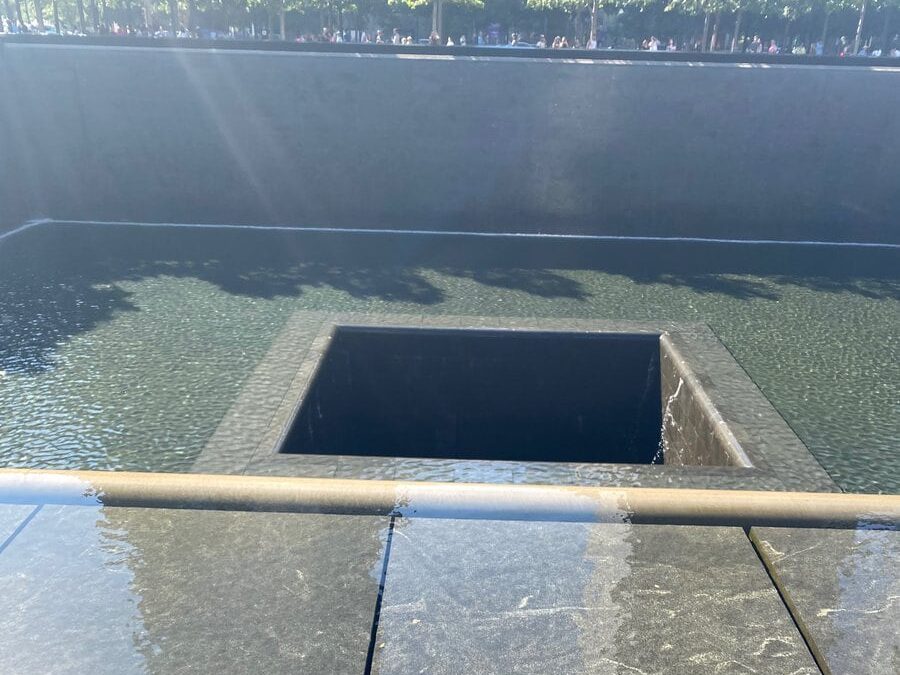 A view of the 9/11 Memorial reflecting pool in New York City, showing water flowing into a central square void, surrounded by smooth stone ledges and a shallow pool reflecting sunlight.